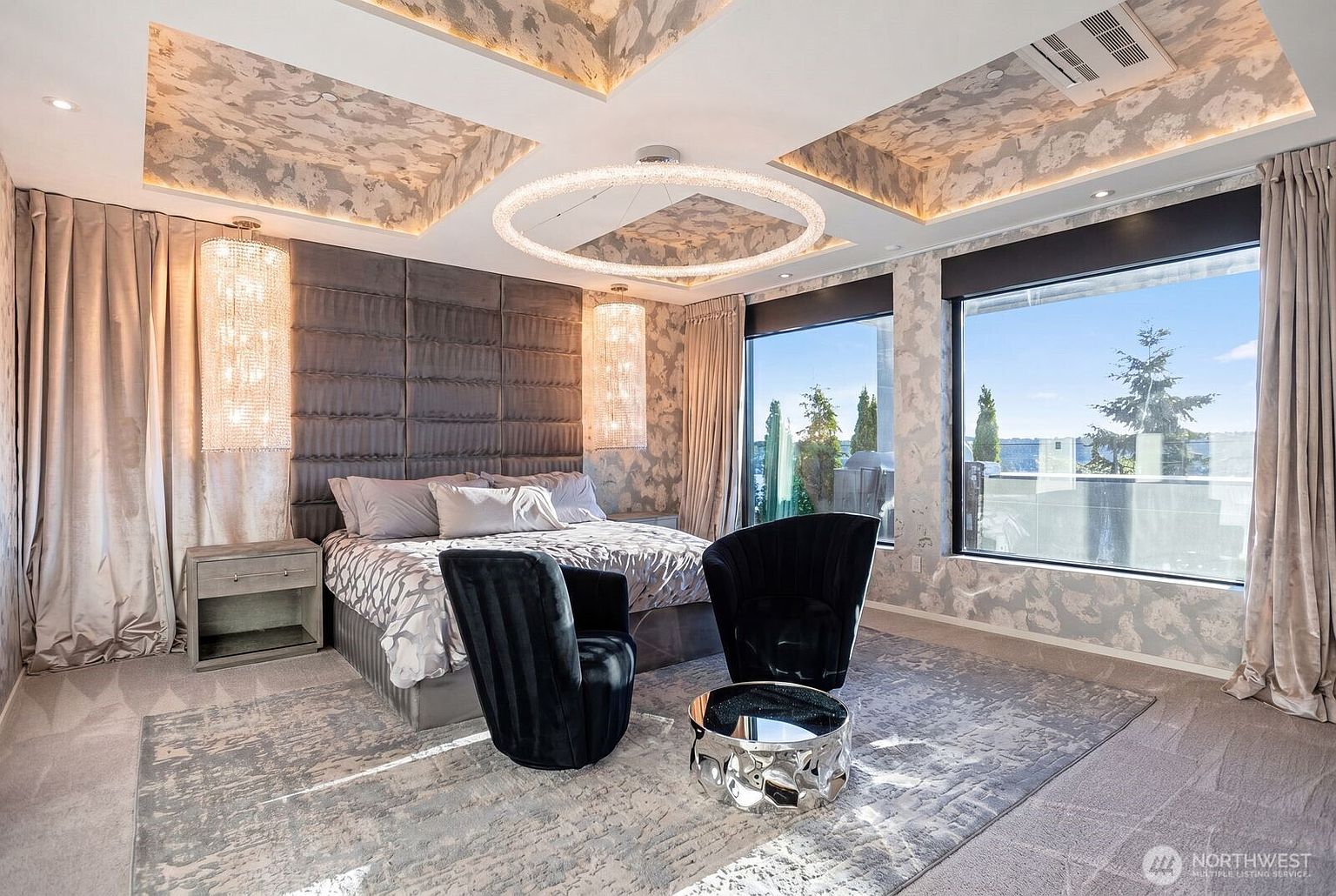 This luxurious primary bedroom features a dramatic upholstered headboard wall and a unique circular chandelier suspended from a recessed, patterned ceiling. Two plush black armchairs are arranged around a reflective, sculptural coffee table, creating a sophisticated seating area in front of large windows that offer expansive outdoor views. The room is finished with elegant drapery, a textured area rug, and crystal-accented bedside lighting, conveying a high-end, contemporary aesthetic.