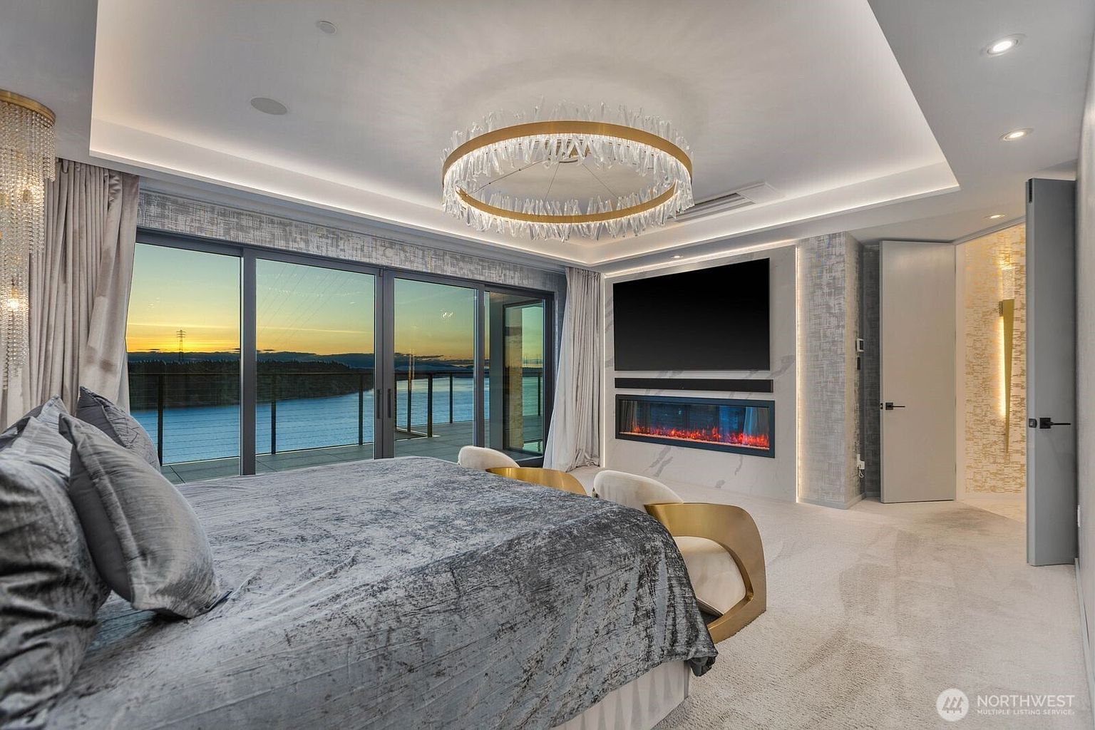 This luxurious primary bedroom features a plush bed with a textured grey coverlet, positioned to face a wall with a built-in television and a linear electric fireplace set into a light-toned stone surround. A striking circular crystal chandelier hangs from the recessed ceiling, while expansive floor-to-ceiling glass doors provide a panoramic view of the water at sunset. The room is finished with soft carpeting and elegant gold-accented furniture, creating a sophisticated and serene atmosphere.