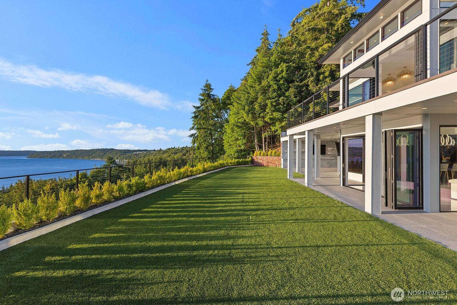 This expansive, manicured lawn overlooks a serene body of water, framed by a lush forest and a clear blue sky. The modern home features a covered patio area with large glass doors and a sleek metal railing, creating a seamless transition between indoor and outdoor living. The perspective captures the vastness of the property, emphasizing the tranquil waterfront setting and the contemporary architectural design.