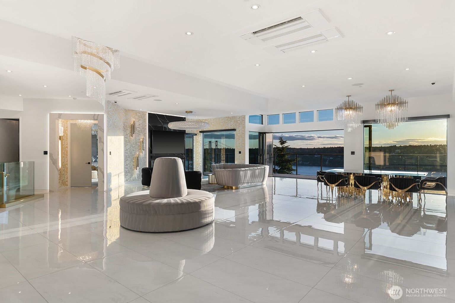 This expansive, open-concept living and dining area features polished light-colored tile flooring and a sophisticated, modern aesthetic. The space is anchored by a unique circular lounge piece in the foreground, with a formal dining set and a plush sofa arrangement positioned toward large glass walls that offer panoramic water views. Elegant crystal chandeliers and recessed lighting illuminate the room, while a dark stone-clad wall serves as a dramatic focal point near the entrance.