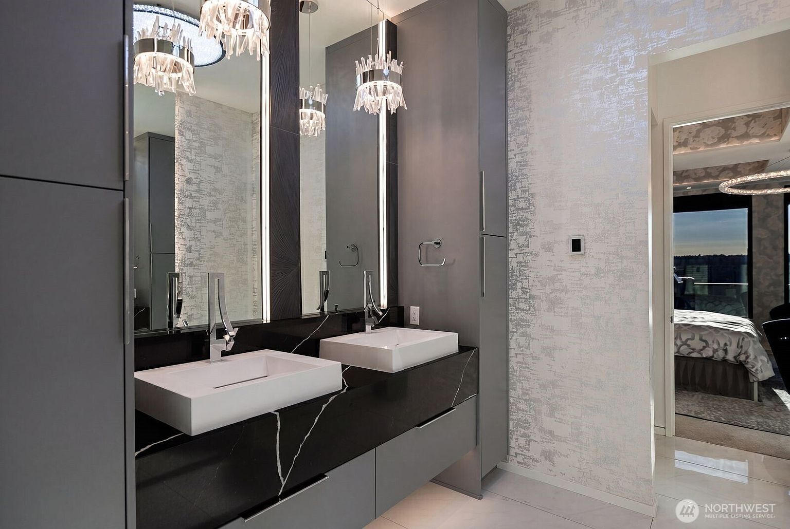 This modern primary bathroom features a sleek dual-vanity setup with rectangular vessel sinks set atop a dark, veined stone countertop. The space is accented by tall, vertical light fixtures flanking the mirrors, crystal-style pendant lights, and textured, metallic-toned wallpaper. A doorway in the background provides a glimpse into a bedroom, emphasizing the open and luxurious flow of the home.