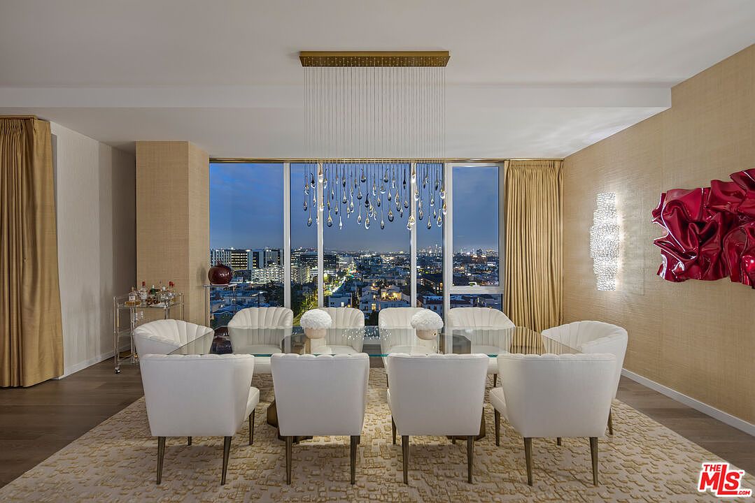 This is an interior shot of a dining room featuring a glass-top table surrounded by white upholstered chairs with wooden legs. A modern chandelier hangs above the table, and large windows offer a city view at dusk. The room is decorated with textured wallpaper and a red abstract sculpture, creating a luxurious and contemporary ambiance.