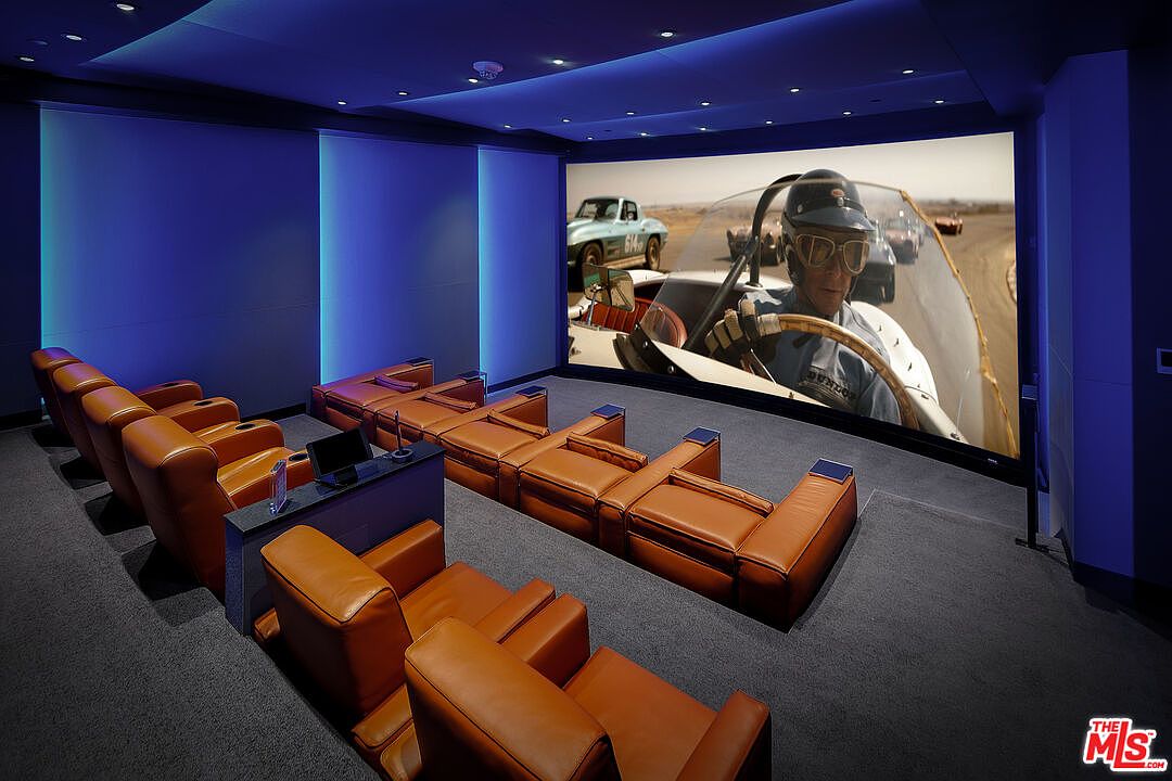This is an interior shot of a home theater, featuring tiered seating upholstered in brown leather. A large screen displays a vintage racing scene, creating a cinematic atmosphere. The room is illuminated with blue ambient lighting, enhancing the immersive experience.