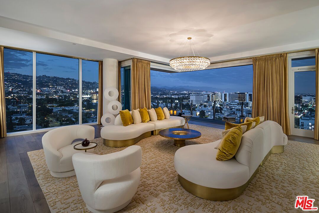 This is a luxurious living room featuring a curved white sofa with gold accents and yellow pillows, complemented by two matching armchairs. A round blue and gold coffee table sits in the center of a patterned rug, while a modern chandelier hangs above. Large windows offer a stunning city view, enhancing the room's elegant and sophisticated ambiance.