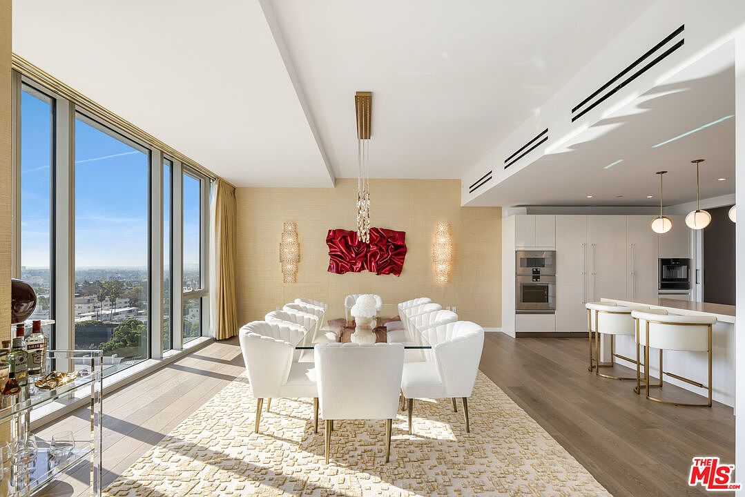 This is a luxurious dining room featuring a glass-top dining table surrounded by white upholstered chairs with gold legs, set on a textured cream-colored rug. The room is illuminated by a modern chandelier and wall sconces, with a large window offering a panoramic city view. The space exudes elegance and sophistication, perfect for upscale dining and entertaining.