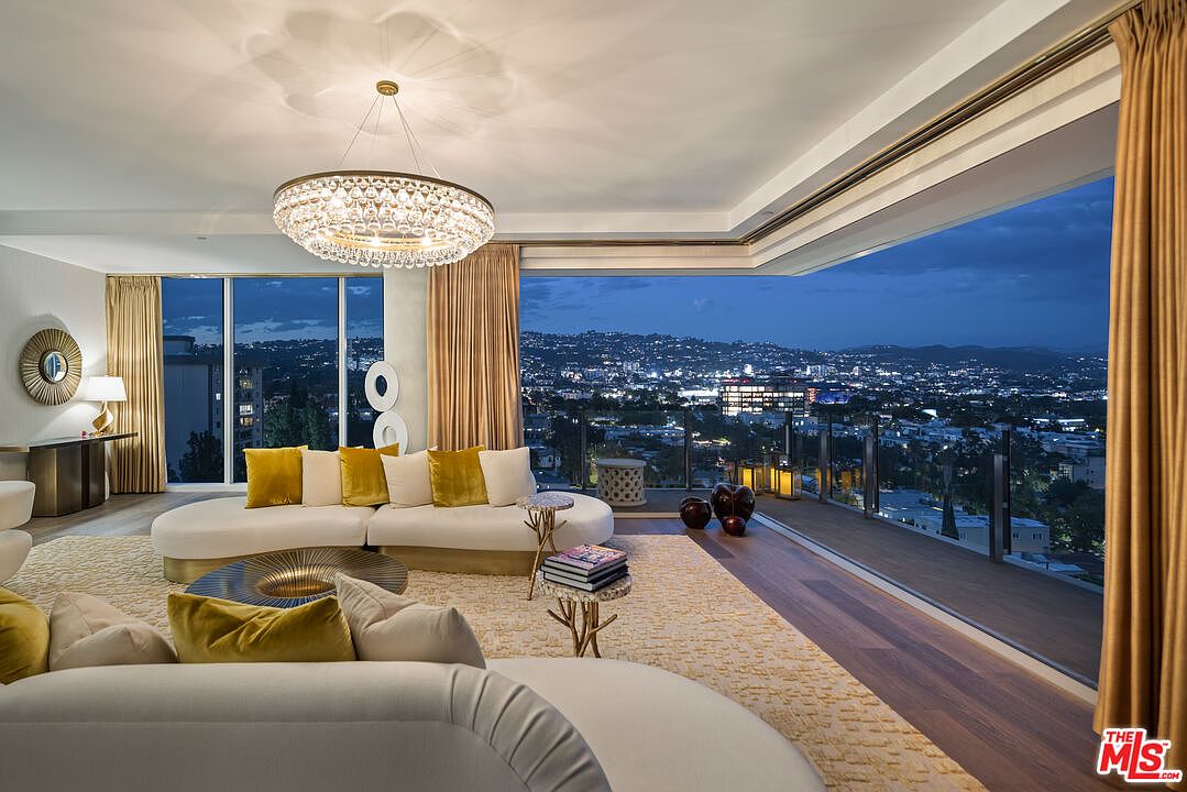 This is a luxurious living room featuring a curved sofa with yellow pillows, a unique coffee table, and a large chandelier. Floor-to-ceiling windows offer a stunning city view at night, and the room is decorated with elegant curtains and modern art pieces. The overall impression is one of sophistication and comfort.