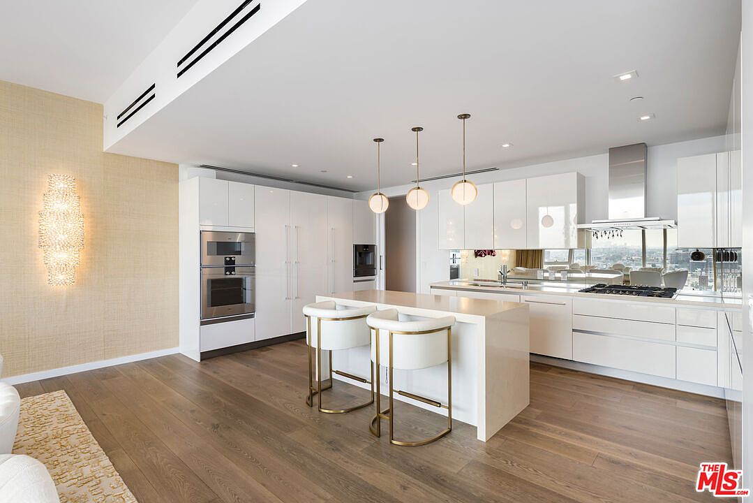 This is a bright, modern kitchen featuring sleek white cabinetry and stainless steel appliances. A central island with bar seating is illuminated by pendant lights, and large windows offer a city view. The hardwood flooring adds warmth to the contemporary design.