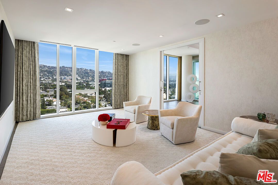This is a bright and airy living room featuring floor-to-ceiling windows that offer panoramic city views. The room is furnished with two plush armchairs, a modern round coffee table, and a comfortable sofa with decorative pillows. The neutral color palette and textured carpet create a sophisticated and inviting atmosphere.