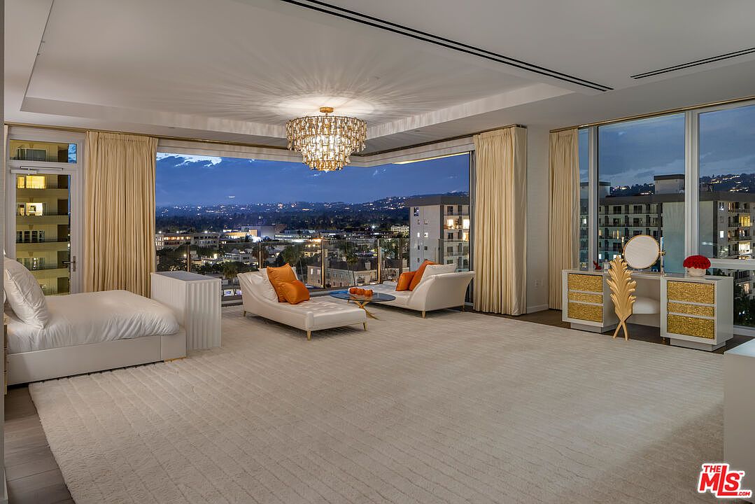 This is a luxurious primary bedroom featuring a large bed, two chaise lounges with orange pillows, and a vanity with a mirror. The room has floor-to-ceiling windows offering a panoramic city view, complemented by elegant curtains and a crystal chandelier. The overall impression is one of opulence and comfort.