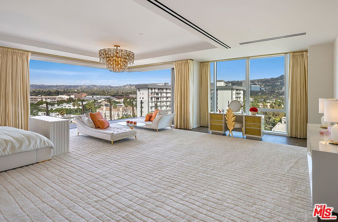 This is a luxurious primary bedroom featuring a spacious layout with a seating area and a vanity. The room boasts floor-to-ceiling windows offering panoramic views, complemented by elegant drapery and a sophisticated chandelier. The overall aesthetic is bright, airy, and upscale, creating a serene and inviting atmosphere.