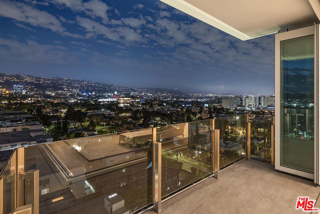 This image showcases a modern balcony with glass railings, offering a stunning panoramic view of the city at night. The balcony features a sleek, minimalist design with a smooth, light-colored floor. The open sliding glass door creates a seamless transition between indoor and outdoor living spaces, highlighting the property's appeal for enjoying city views.