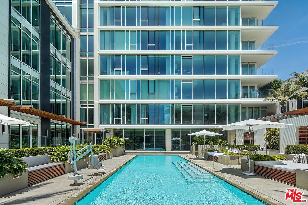 This image showcases a luxurious outdoor pool area, featuring a rectangular pool with clear blue water and steps leading into it. The pool is surrounded by modern lounge seating, umbrellas, and lush greenery, creating a serene and upscale atmosphere. The backdrop includes a contemporary building with large glass windows, suggesting a high-end residential complex.
