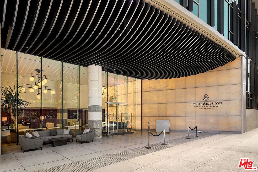 The image showcases the grand entryway of the Four Seasons Private Residences in Los Angeles. A modern, curved canopy shelters the entrance, which features floor-to-ceiling glass walls and a prominent marble-clad wall with the building's logo. The space is furnished with contemporary seating, creating an inviting and luxurious arrival experience.