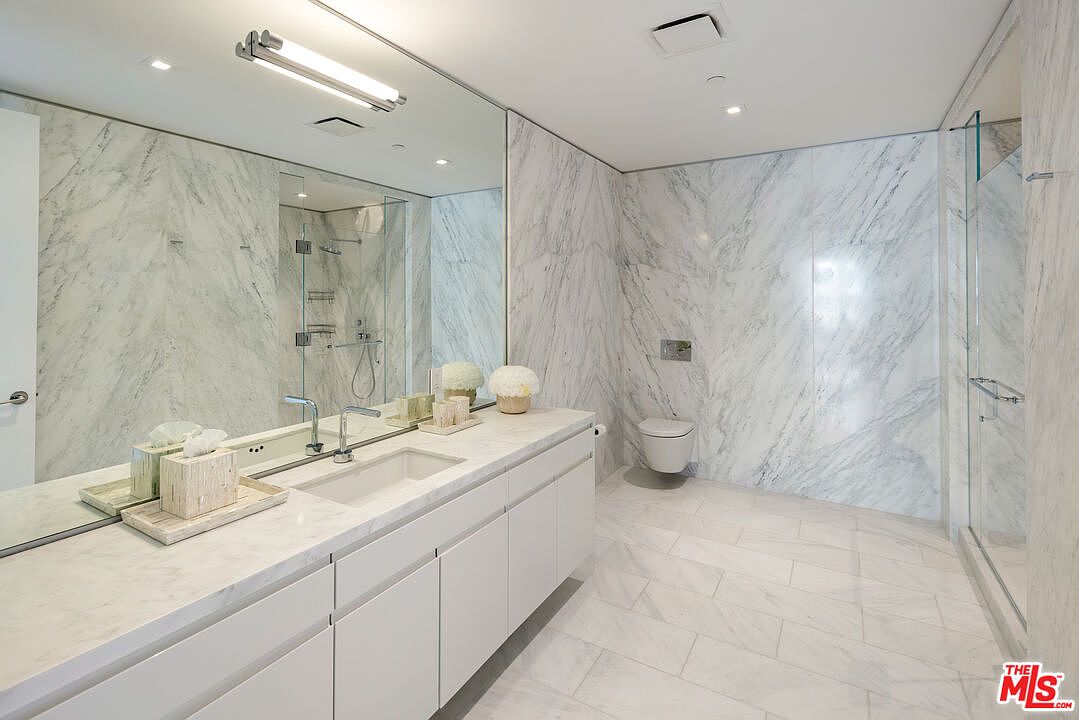 This is a luxurious primary bathroom featuring marble walls and flooring, creating a seamless and elegant design. A long, white vanity with integrated sink and minimalist cabinetry stretches across the wall, complemented by a large mirror and modern lighting fixture. A glass-enclosed shower is visible, adding to the spa-like atmosphere of the space.