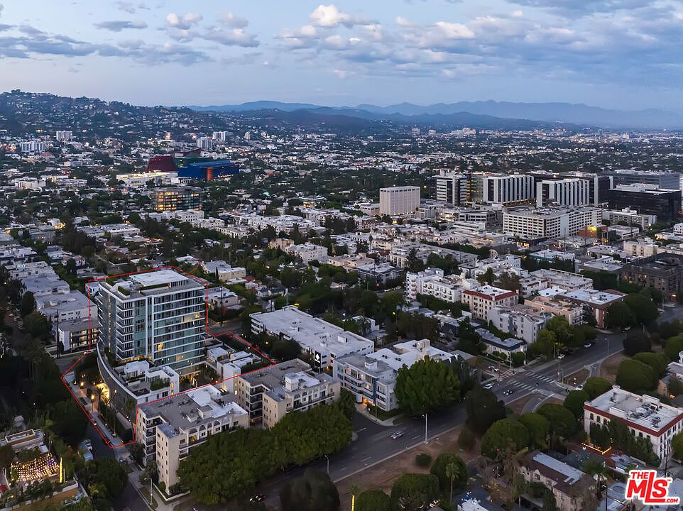 This aerial view showcases a modern residential building with a glass facade, surrounded by lush greenery and other low-rise buildings. The property is outlined in red, emphasizing its boundaries within the urban landscape. The image captures a sense of upscale living within a vibrant city setting, with mountains visible in the distance.
