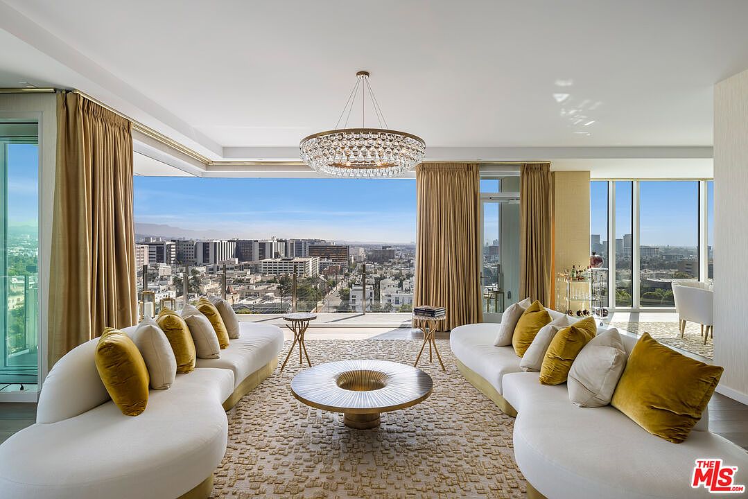 This is a luxurious living room featuring two curved white sofas with gold accents and plush pillows, facing each other across a textured beige rug and a unique round coffee table. A stunning crystal chandelier hangs from the ceiling, and floor-to-ceiling windows offer panoramic city views, creating a bright and airy atmosphere. The room is elegantly styled with gold curtains and side tables, exuding sophistication and comfort.