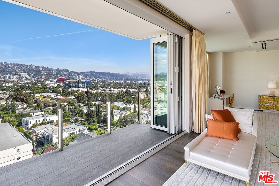 This image showcases a luxurious living room with a stunning view of the city skyline from a spacious balcony. The interior features a stylish chaise lounge with decorative pillows, complemented by elegant curtains and a modern vanity area. The open balcony with glass railings enhances the sense of space and provides an inviting outdoor extension to the living area.