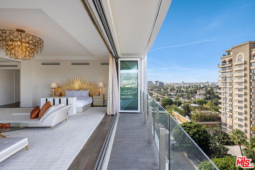 This primary bedroom boasts an open layout with a luxurious seating area and a private balcony offering stunning city views. The room features a unique sunburst headboard, a dazzling chandelier, and a neutral color palette, creating an elegant and serene atmosphere. The seamless transition to the outdoor balcony enhances the sense of spaciousness and sophistication.