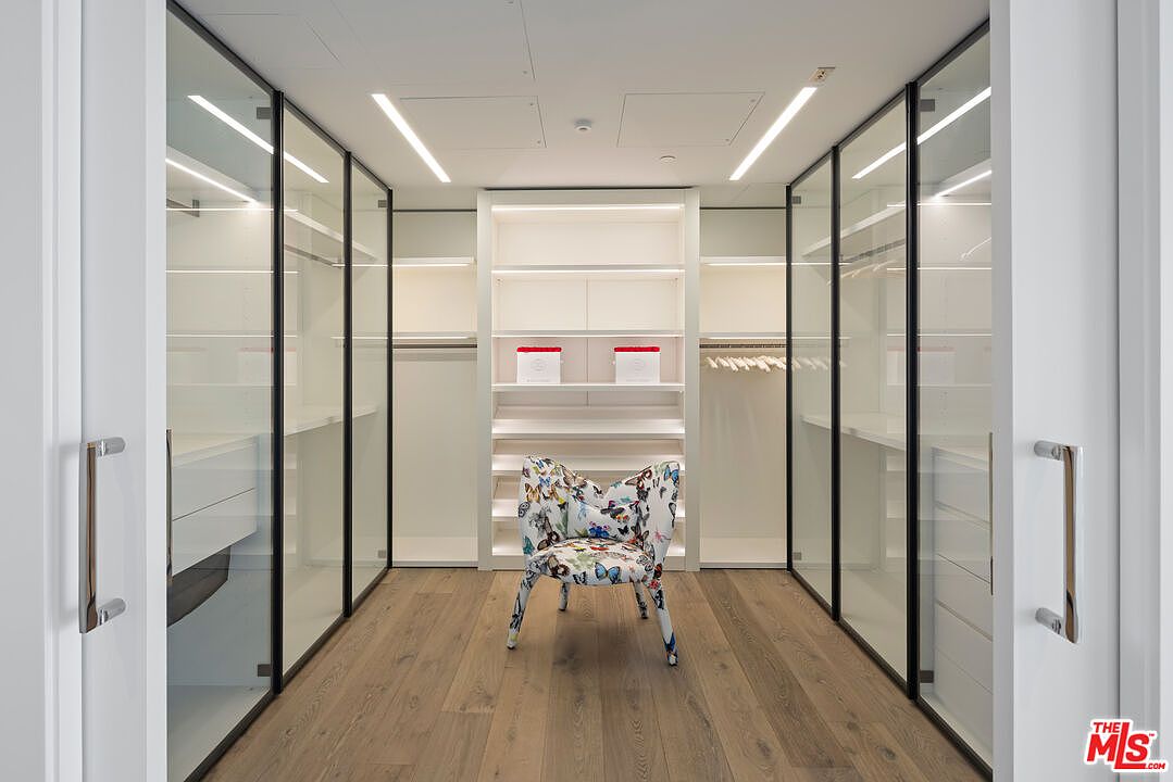 This is a well-organized walk-in closet featuring glass-fronted wardrobes with black frames, white shelving, and a central shelving unit. A patterned chair sits in the middle of the room, adding a touch of personality. The closet is illuminated by modern linear lighting, and the hardwood flooring adds warmth to the space.