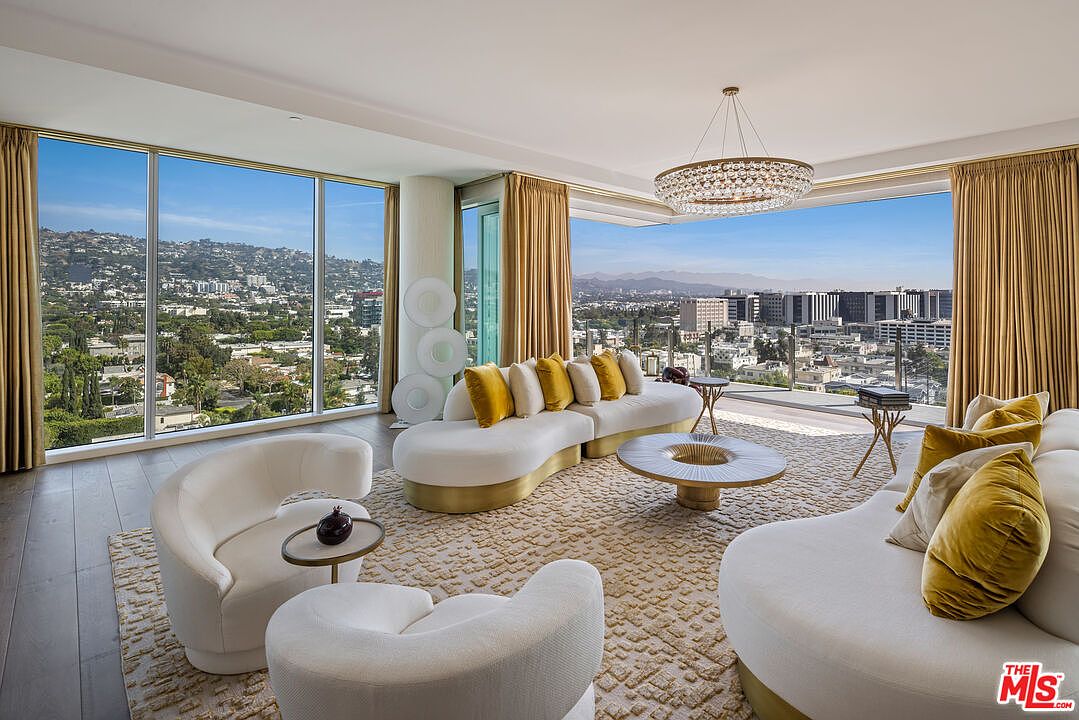 This is a luxurious living room featuring a curved white sofa with gold accents and yellow pillows, complemented by matching armchairs and a unique circular coffee table. Large windows offer panoramic city views, enhanced by elegant gold curtains and a modern chandelier. The overall impression is one of sophisticated elegance and comfort.