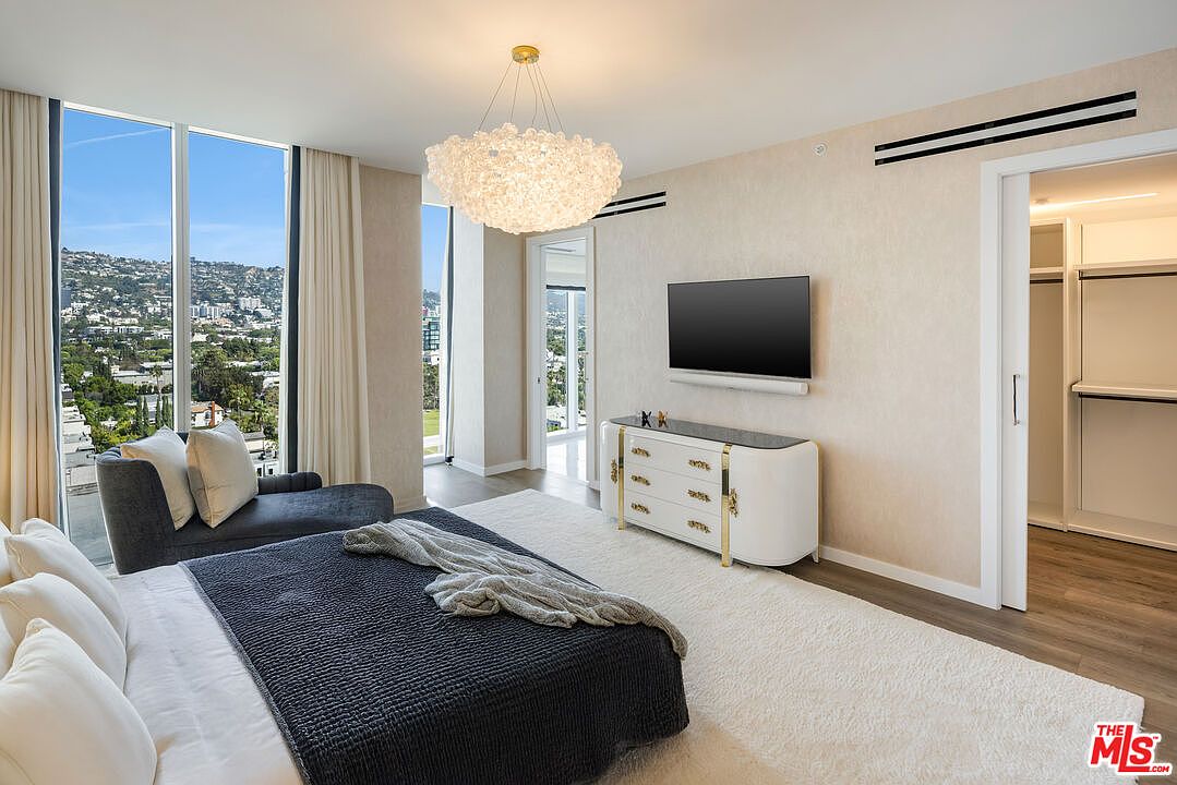 This is a luxurious primary bedroom featuring a large bed with a dark gray comforter and white pillows, a gray chaise lounge by a large window with a city view, and a white dresser with gold accents beneath a wall-mounted TV. The room is decorated in neutral tones with a plush white rug, creating a serene and upscale atmosphere. The perspective is from the foot of the bed, looking towards the window and dresser.