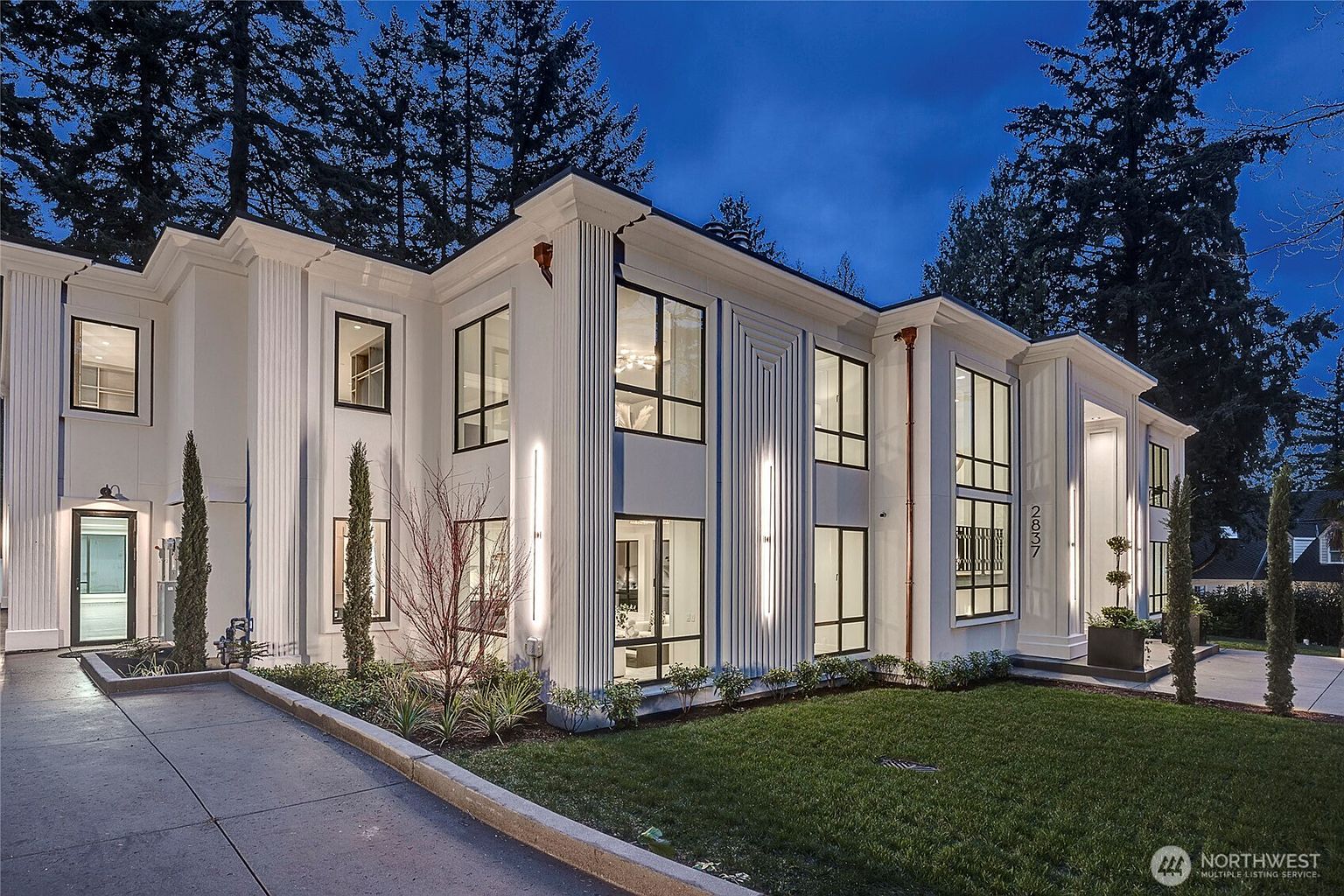 This striking modern luxury home features a clean, white facade with dramatic vertical architectural detailing and large, floor-to-ceiling windows. The exterior is illuminated by sleek, integrated wall lighting, creating a sophisticated and inviting atmosphere at dusk. A manicured lawn and structured landscaping frame the property, highlighting its contemporary design and curb appeal.