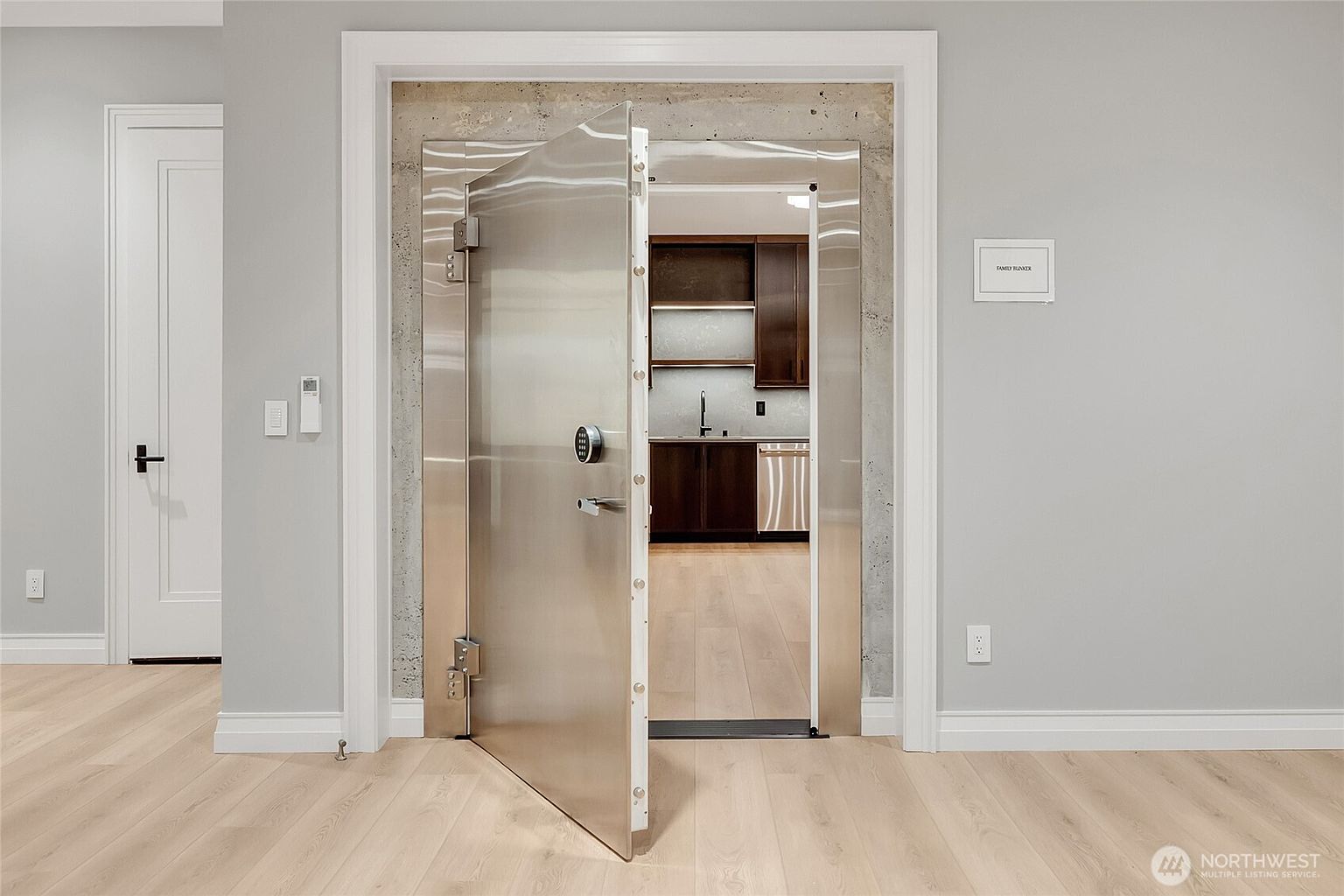 This image features a heavy-duty, industrial-style stainless steel vault door serving as the entrance to a secure room, labeled as a 'Family Bunker'. The door is set within a raw concrete frame, contrasting with the surrounding light-colored walls and hardwood flooring, while a glimpse into the interior reveals a modern kitchen setup. The perspective is a straight-on, eye-level shot that emphasizes the imposing nature of the security door against the otherwise residential interior design.