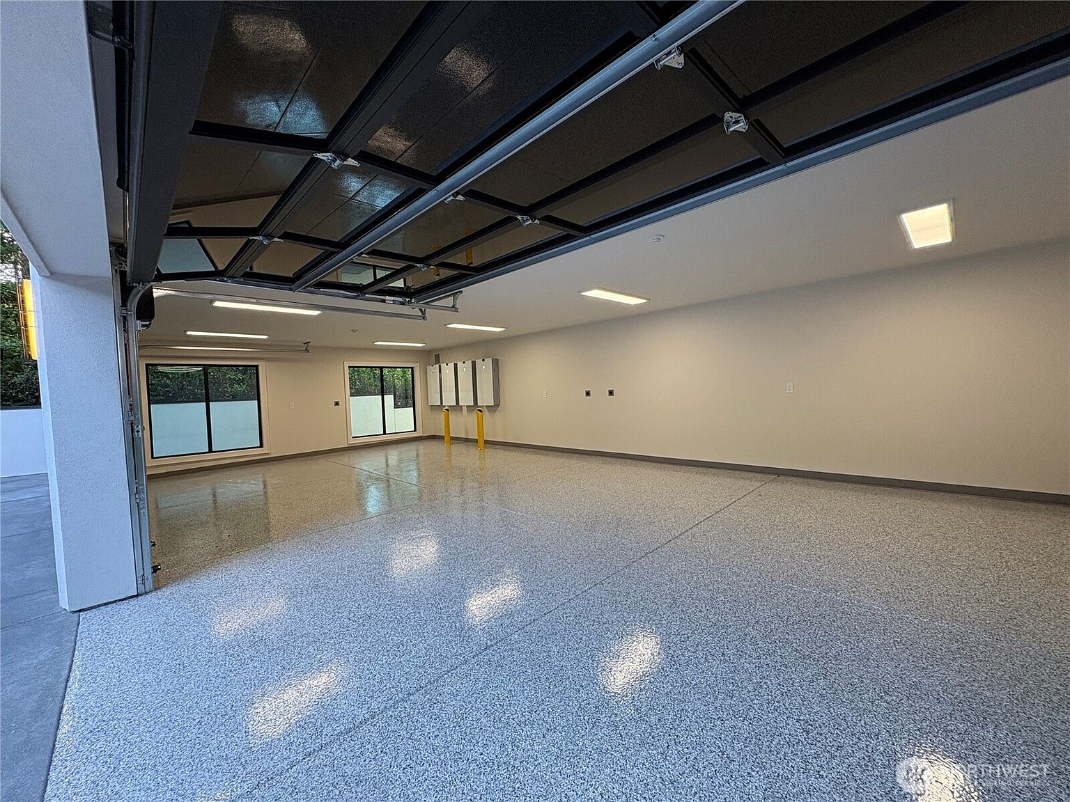 This spacious, modern garage features a pristine, speckled epoxy floor and bright, clean white walls. The perspective is from inside looking out, showcasing the open overhead door, multiple linear LED light fixtures, and two windows that provide natural light. The space is uncluttered and well-maintained, offering a versatile area for parking or a workshop.