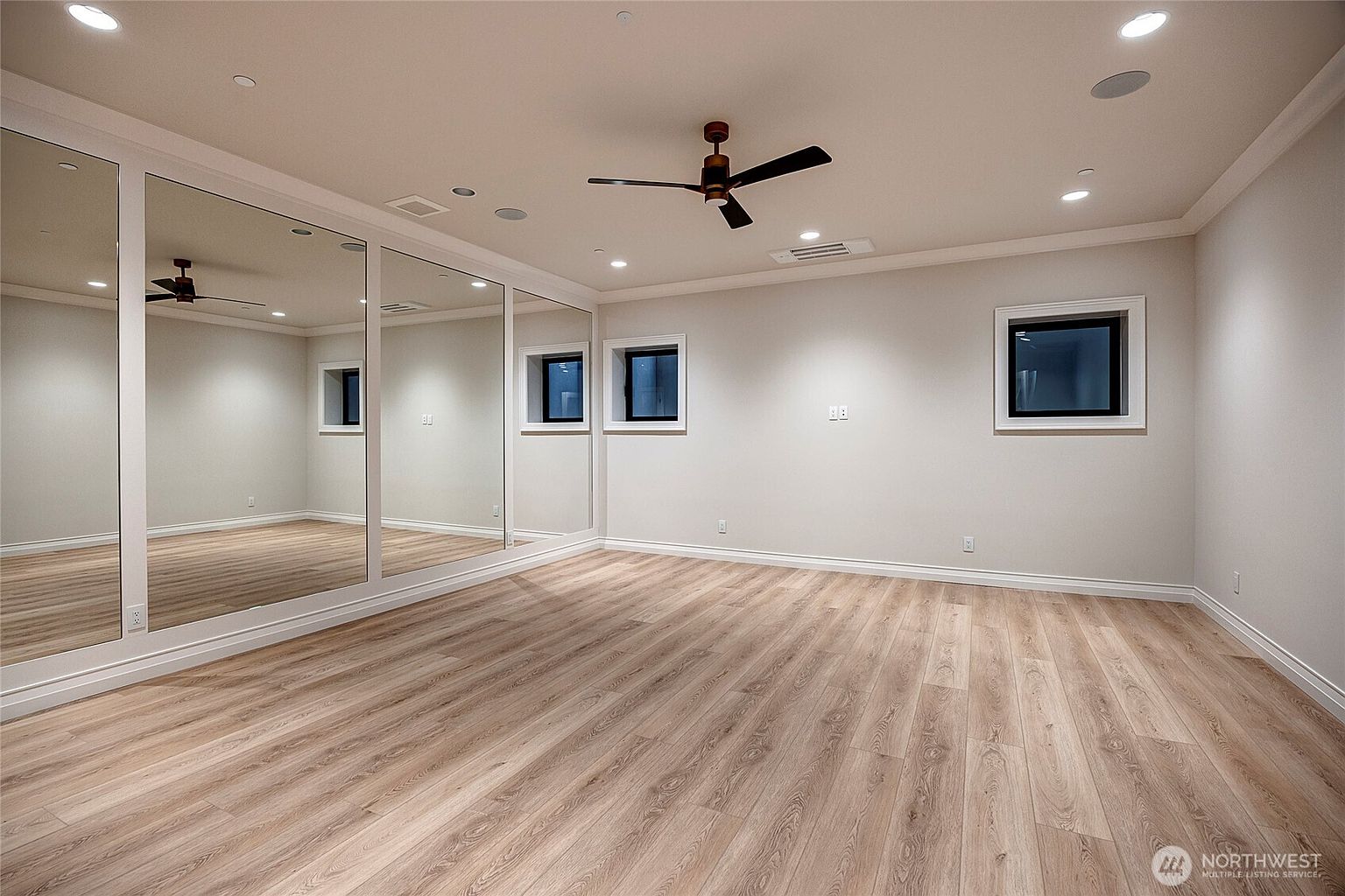 This spacious, minimalist room features light wood-look flooring and a wall of floor-to-ceiling mirrors, making it an ideal space for a home gym or dance studio. The room is illuminated by recessed lighting and a central ceiling fan, with several small windows providing natural light. The neutral color palette and clean lines create a versatile, open environment.