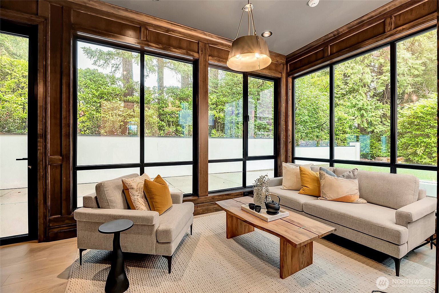 This elegant sunroom features floor-to-ceiling windows framed in rich, dark wood paneling, creating a seamless connection to the lush greenery outside. The space is furnished with a comfortable light-gray sofa and matching armchair, accented by warm mustard-toned pillows and a natural wood coffee table. A modern, oversized pendant light hangs from the ceiling, casting a soft glow over the neutral-toned area rug, resulting in a sophisticated and tranquil atmosphere.