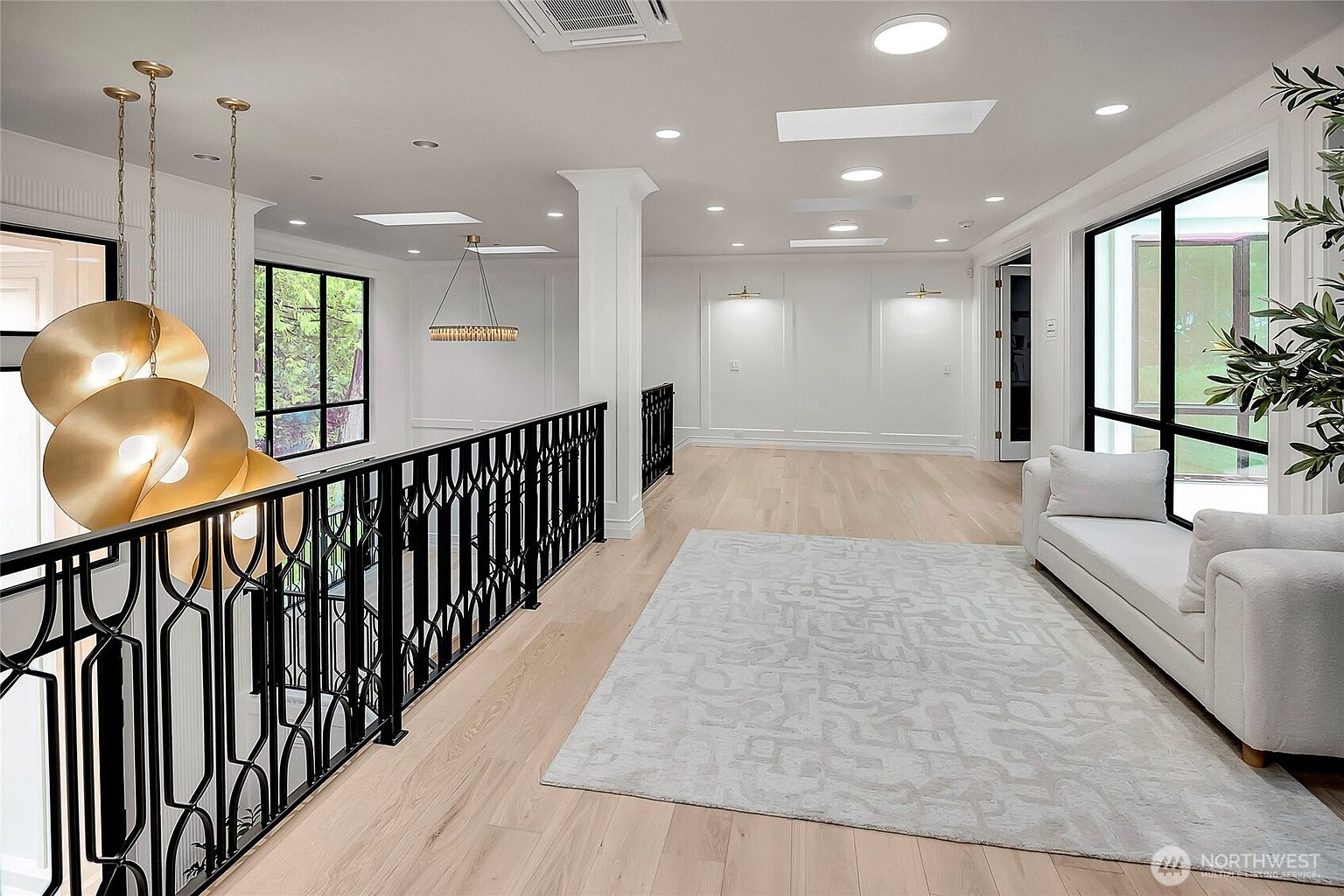 This elegant, light-filled landing area features light hardwood flooring, a modern white sofa, and a sophisticated black metal railing overlooking the lower level. The space is accented by a striking gold pendant light fixture and recessed lighting, creating a bright and airy atmosphere. The clean lines and minimalist decor emphasize a contemporary, high-end aesthetic.