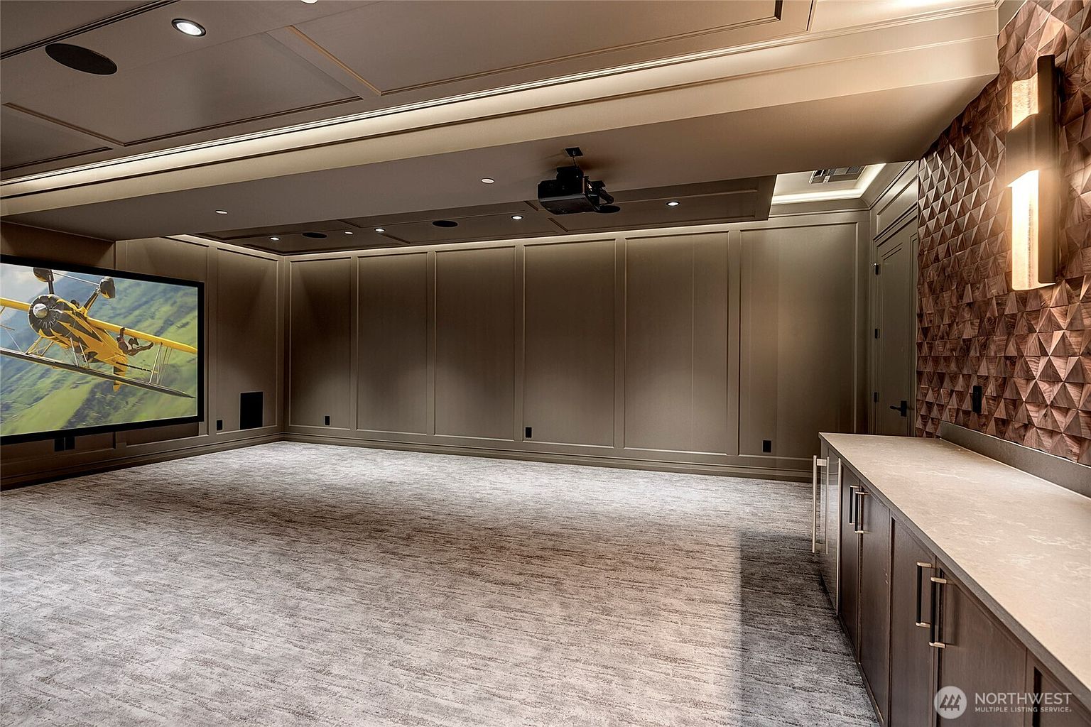 This sophisticated home theater features a large projection screen displaying a vintage airplane, complemented by elegant wall paneling and a textured, geometric accent wall. A sleek, dark-toned wet bar with a stone countertop runs along the right side, providing a functional space for refreshments. The room is finished with plush carpeting and recessed lighting, creating a luxurious and immersive cinematic environment.