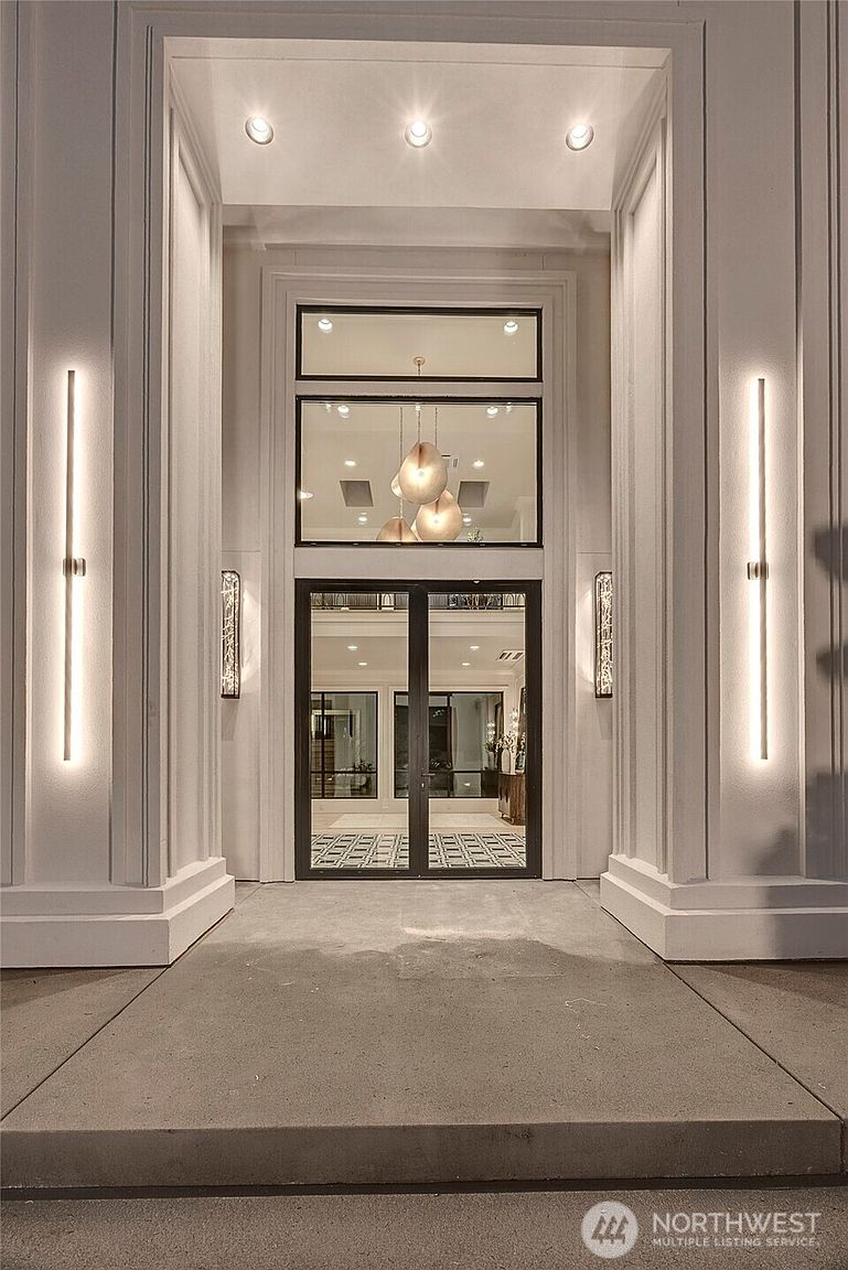 This striking front entryway features a grand, symmetrical design with tall, vertical LED light fixtures flanking a modern double-door entrance. The architectural style is contemporary and sophisticated, highlighted by recessed ceiling lights and a large transom window that reveals a glimpse of an elegant interior chandelier. The clean lines and neutral color palette create a welcoming and luxurious first impression for the home.