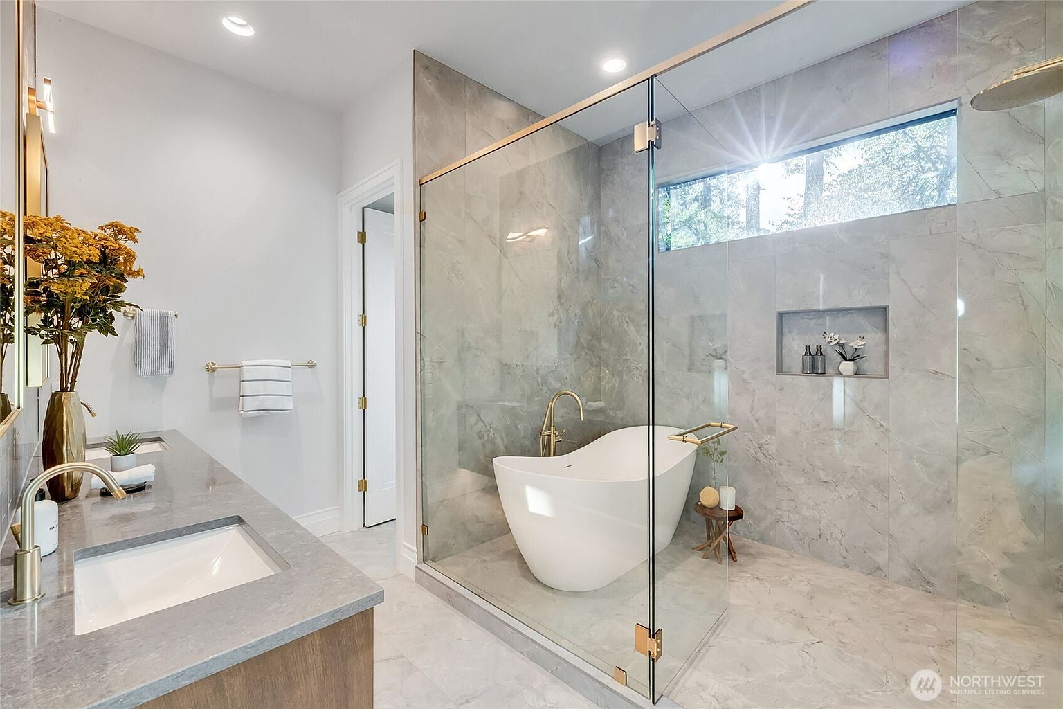 This luxurious primary bathroom features a sleek, glass-enclosed wet room containing a modern freestanding soaking tub and a rainfall shower. The space is elegantly finished with large-format marble-look tile, warm gold hardware, and a double vanity with a grey stone countertop. The overall aesthetic is sophisticated and spa-like, emphasizing high-end materials and clean, contemporary lines.