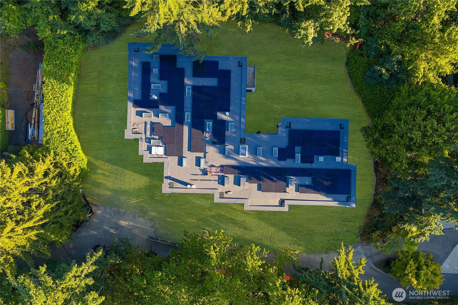 This high-angle aerial view captures a sprawling, flat-roofed residence nestled within a lush, tree-lined property. The roof features extensive solar panel installations and multiple skylights, set against a backdrop of vibrant green lawn and dense surrounding foliage. The perspective emphasizes the unique, expansive footprint of the home and its secluded, park-like setting.