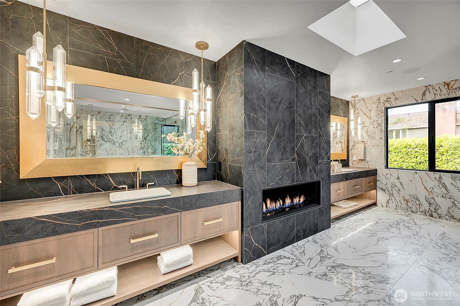 This luxurious primary bathroom features a striking dual-vanity setup with dark, gold-veined marble countertops and custom wood cabinetry. A central fireplace encased in matching dark marble serves as a dramatic focal point, while a large skylight and elegant crystal pendant lighting enhance the sophisticated, high-end aesthetic. The space is finished with white marble flooring and expansive mirrors, creating a bright, opulent atmosphere.
