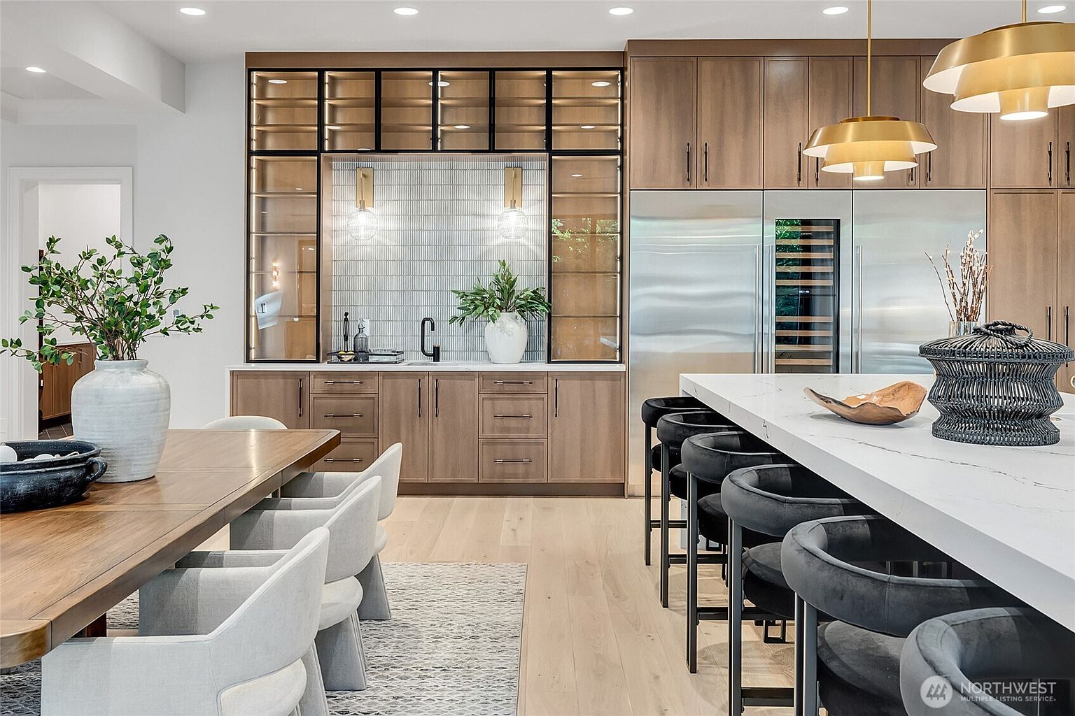 This sophisticated, modern kitchen features a seamless blend of warm wood cabinetry and sleek, contemporary design elements. A large white quartz island with bar seating serves as the centerpiece, complemented by a dedicated wet bar area with glass-front cabinetry and elegant tile backsplash. The space is bathed in warm, ambient lighting from stylish pendant fixtures, creating an inviting and luxurious atmosphere perfect for entertaining.