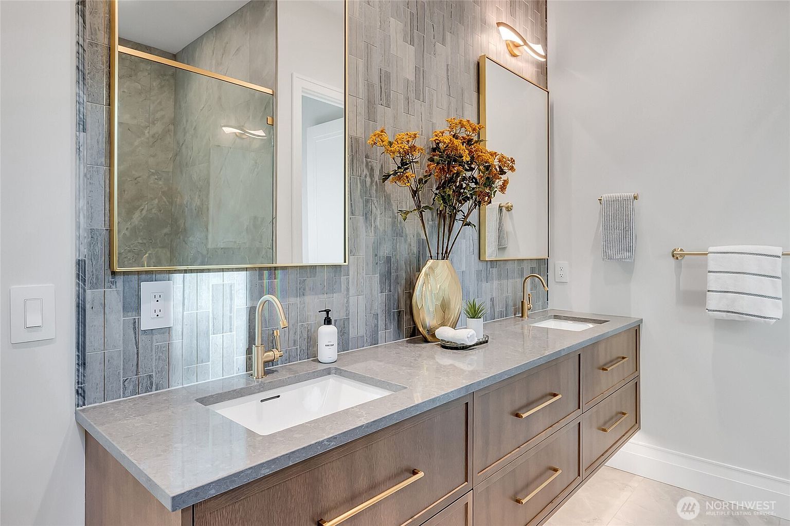 This modern primary bathroom features a sleek double vanity with a gray quartz countertop, warm wood cabinetry, and elegant gold hardware. The wall is adorned with vertical gray textured tiles, complemented by two gold-framed mirrors and contemporary wall sconces. The overall aesthetic is sophisticated and luxurious, creating a spa-like atmosphere.