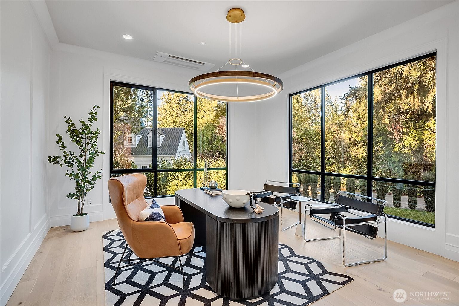 This sophisticated home office features a modern, dark-toned desk as the centerpiece, complemented by a tan leather wingback chair and two sleek black leather guest chairs. The room is bathed in natural light from expansive floor-to-ceiling windows that offer a serene view of the surrounding greenery, while a geometric area rug and a contemporary circular chandelier add a touch of artistic flair. The minimalist white walls and light hardwood floors create a bright, professional, and tranquil workspace.