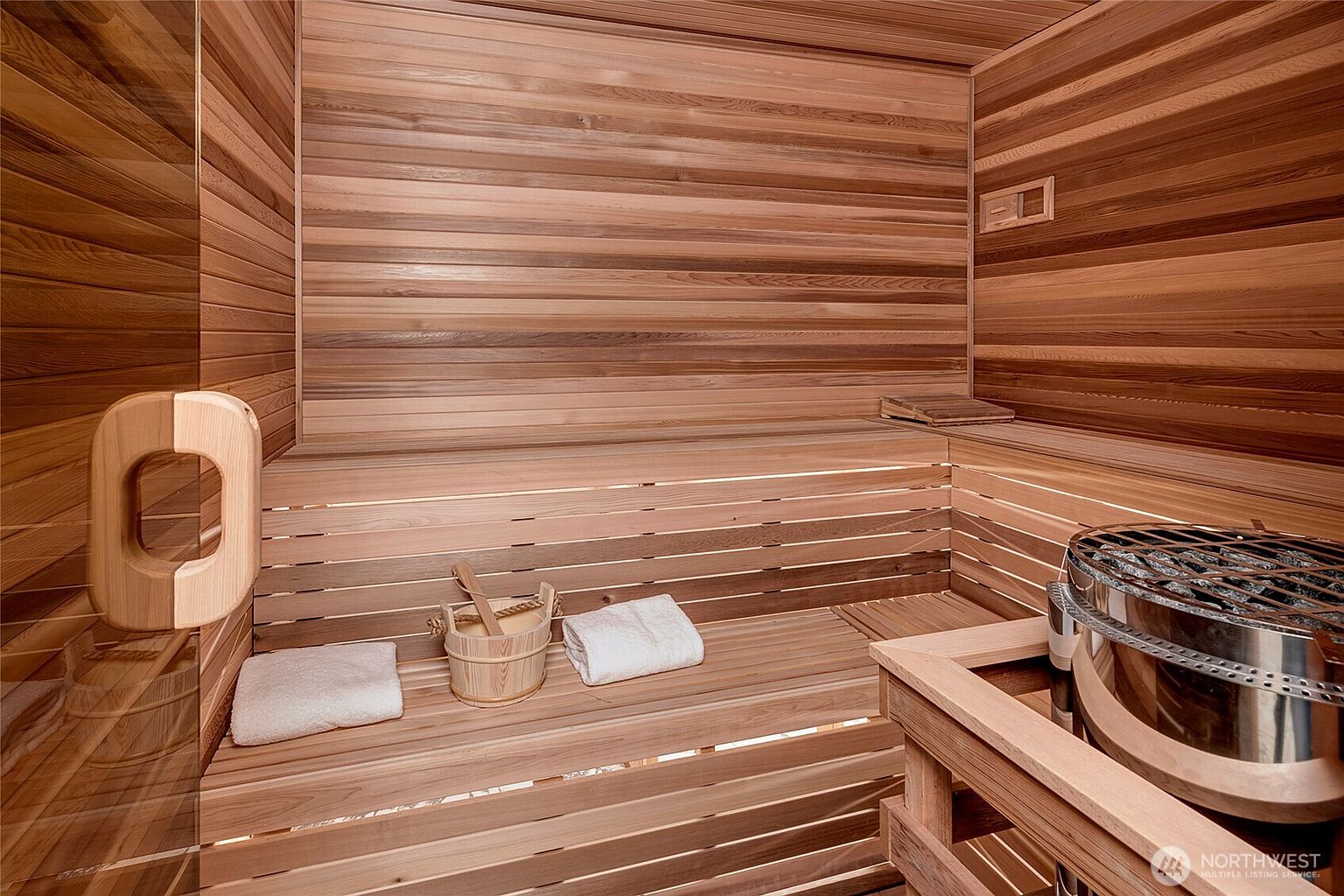 This image showcases a traditional cedar-lined sauna, featuring tiered wooden benches and a prominent metal heater unit in the foreground. A wooden bucket and ladle sit on the bench alongside folded white towels, emphasizing a spa-like, relaxing atmosphere. The warm, natural wood tones and clean lines create an inviting and luxurious wellness space.