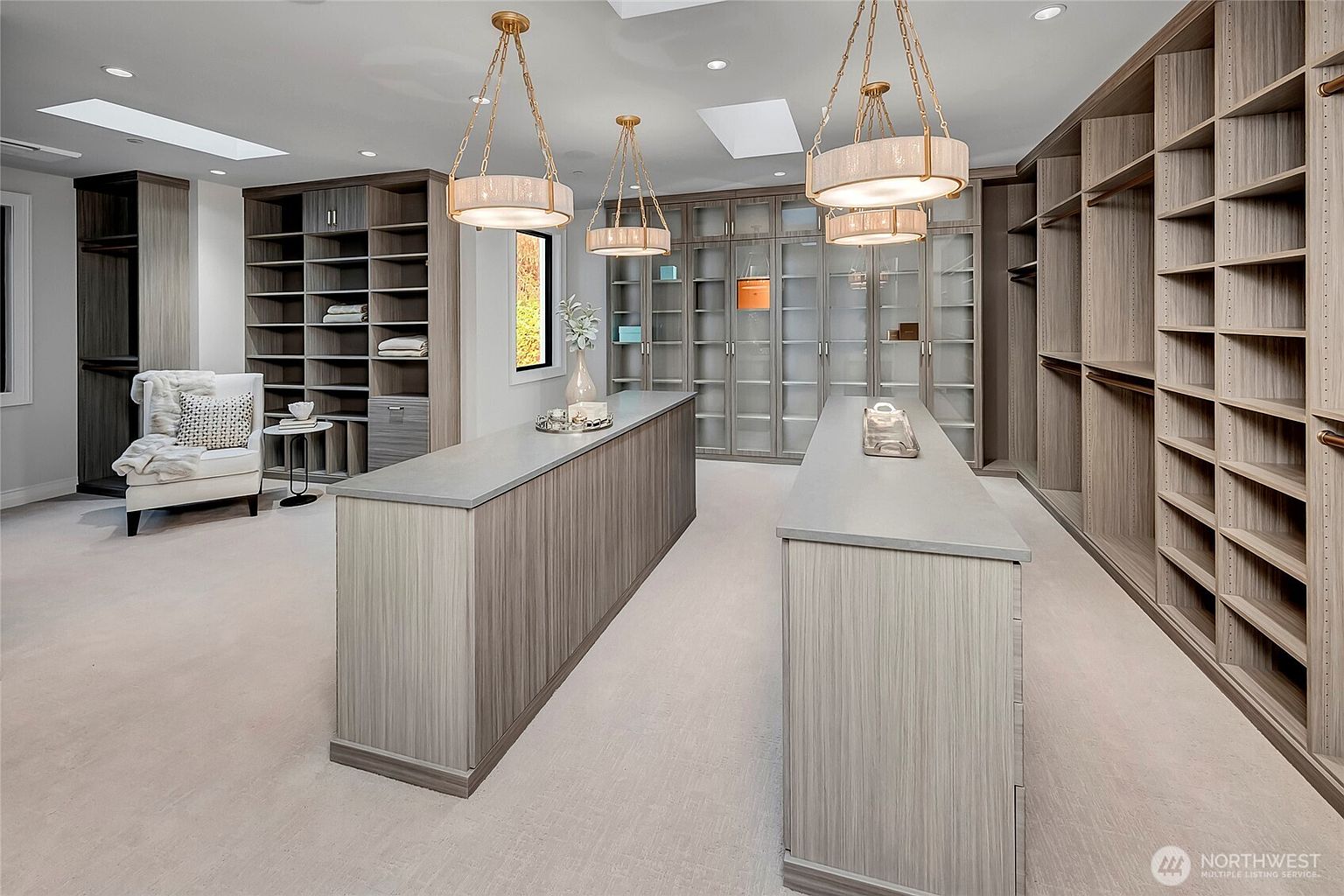 This expansive, high-end walk-in closet features custom light-wood cabinetry, two central islands with stone countertops, and elegant gold-toned pendant lighting. The room is designed with a mix of open shelving, glass-fronted display cabinets, and a cozy seating area, all illuminated by recessed lighting and skylights. The overall aesthetic is sophisticated, organized, and luxurious, providing a bright and airy atmosphere.