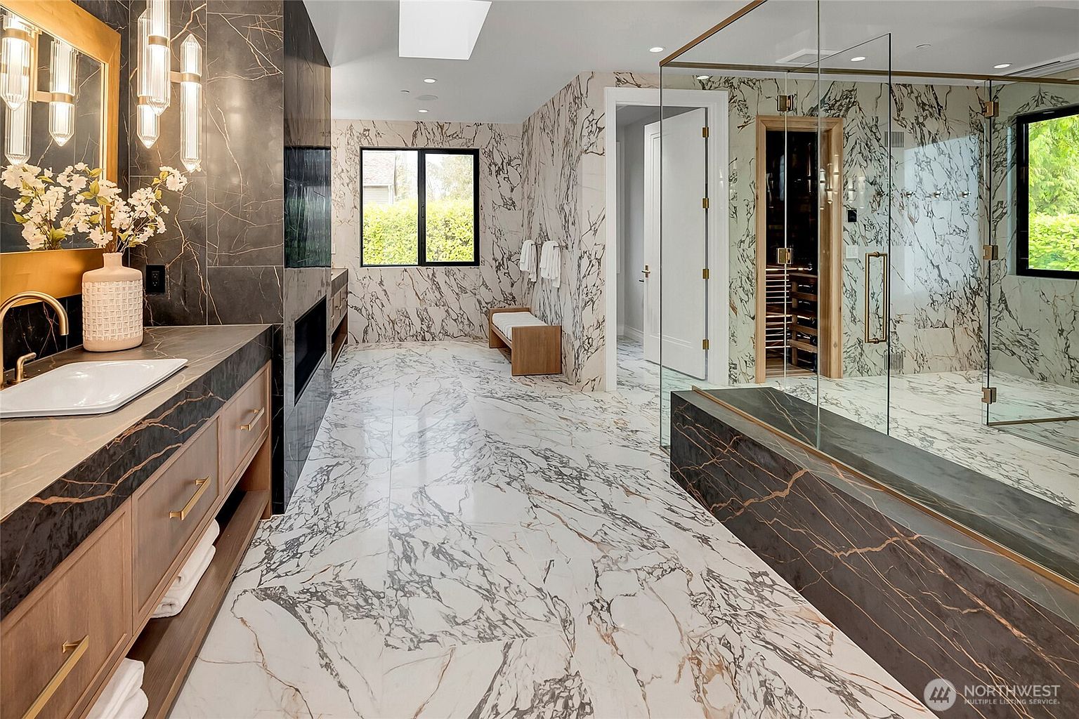 This luxurious primary bathroom features striking, high-contrast marble surfaces throughout, including the flooring, walls, and a custom vanity. The space is equipped with a glass-enclosed shower and a built-in sauna, creating a spa-like atmosphere. The perspective captures the expansive layout, highlighting the sophisticated blend of warm wood cabinetry and dramatic veined stone.