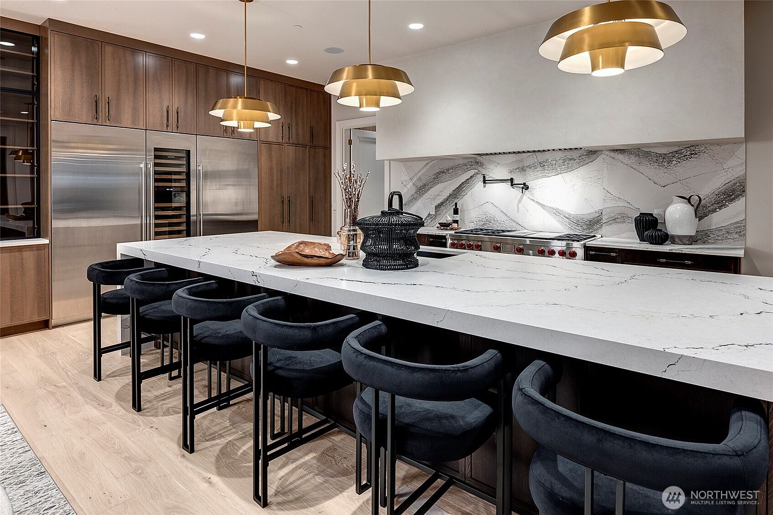 This sophisticated, modern kitchen features a massive waterfall-edge quartz island with dark velvet bar stools, serving as the centerpiece of the room. The space is defined by warm wood cabinetry, high-end stainless steel appliances, and a striking marble slab backsplash with a pot filler faucet. Two large, tiered gold pendant lights hang above the island, creating a luxurious and inviting atmosphere.