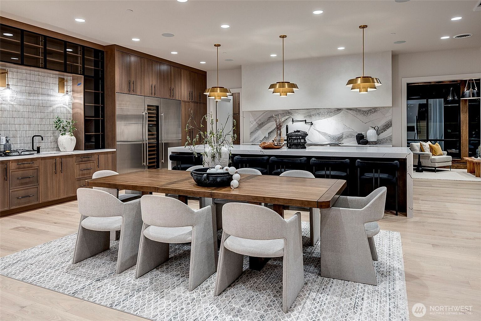 This sophisticated, open-concept kitchen and dining area showcases a seamless blend of modern luxury and warm, natural textures. The space features a large waterfall-edge island with bar seating, complemented by rich wood cabinetry, a striking marble backsplash, and elegant gold pendant lighting. A spacious dining table with contemporary upholstered chairs sits in the foreground, creating a perfect setting for both casual meals and formal entertaining.
