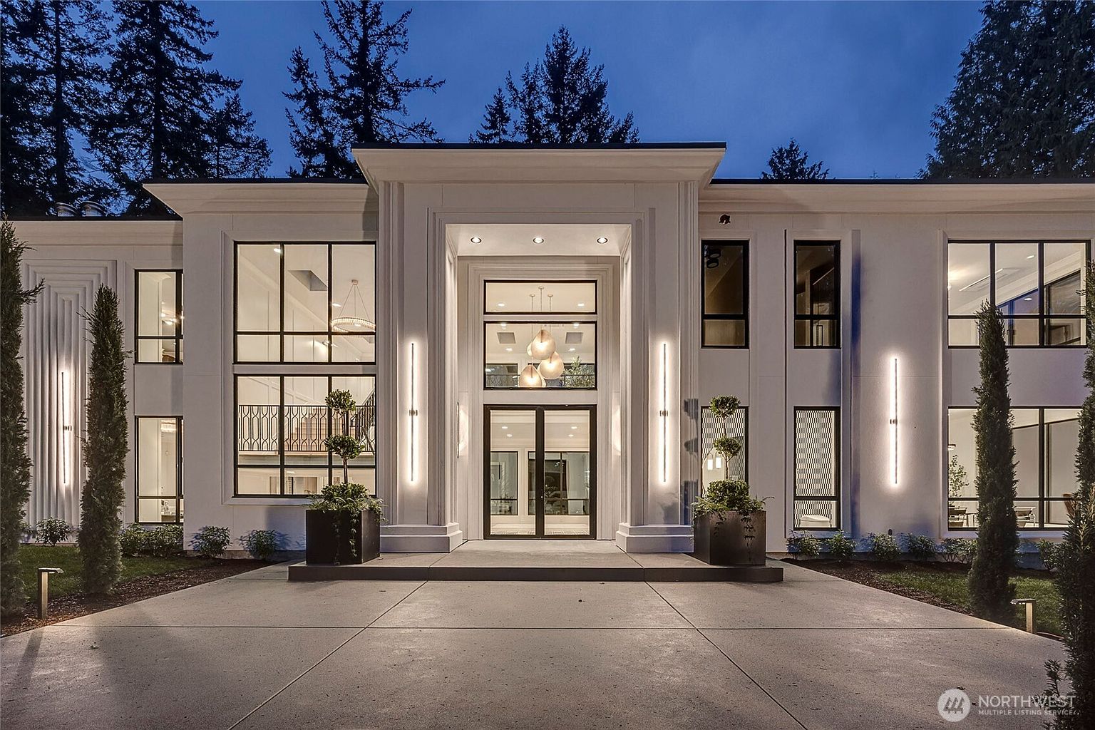 This striking modern estate features a symmetrical, grand facade with clean white lines and expansive floor-to-ceiling windows. The central entryway is highlighted by a towering portico, vertical accent lighting, and a sophisticated glass-paneled door, creating an imposing and luxurious first impression. The manicured landscaping, including tall, slender cypress trees and structured planters, perfectly complements the contemporary architectural design.