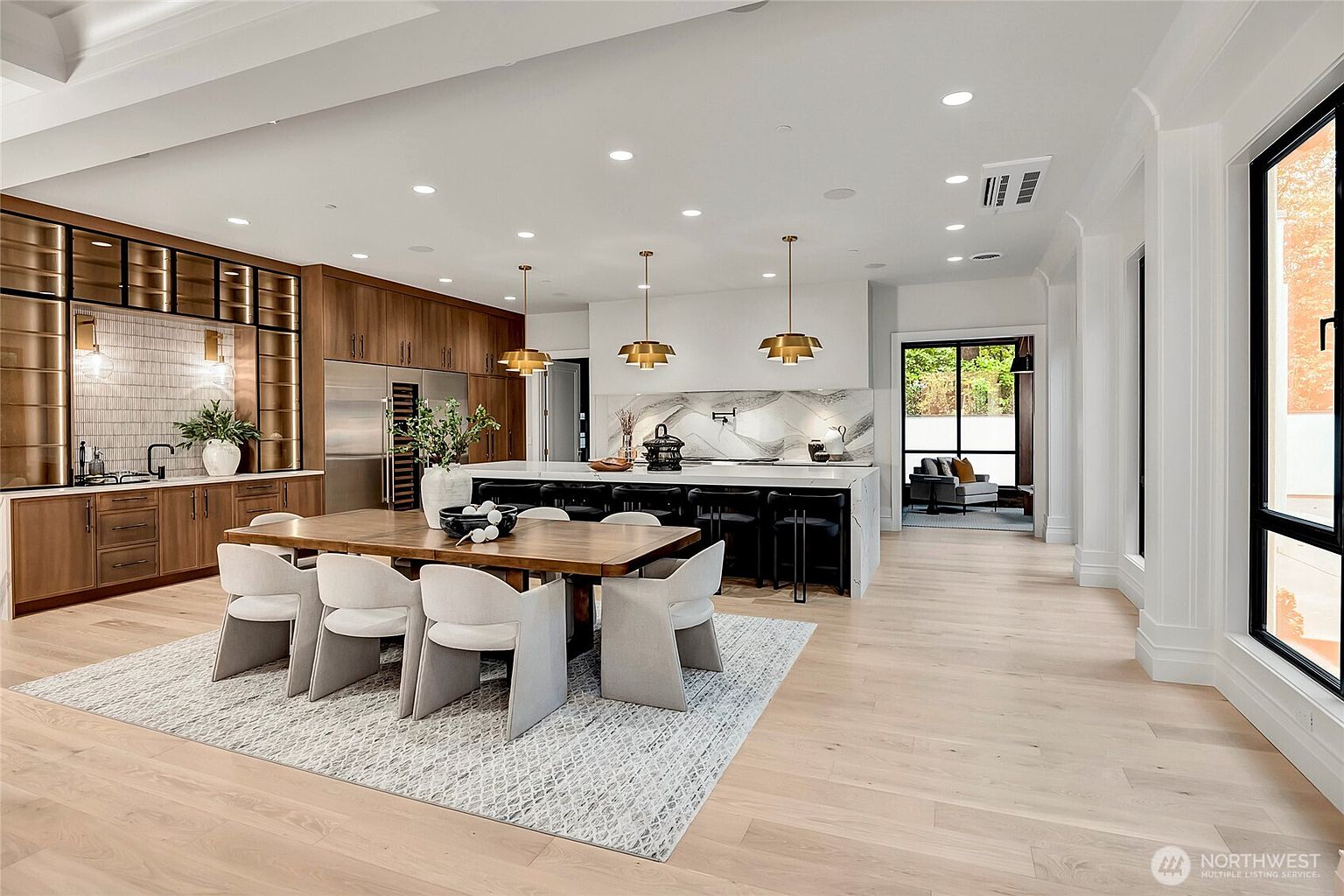 This expansive, modern open-concept kitchen and dining area features light hardwood flooring, sleek wood cabinetry, and a large marble-topped island with black bar stools. A sophisticated dining table with contemporary upholstered chairs sits on a textured rug, while three gold pendant lights provide elegant illumination over the island. The space is bright and airy, with large windows and a seamless transition to an adjacent lounge area, creating a luxurious and inviting atmosphere.