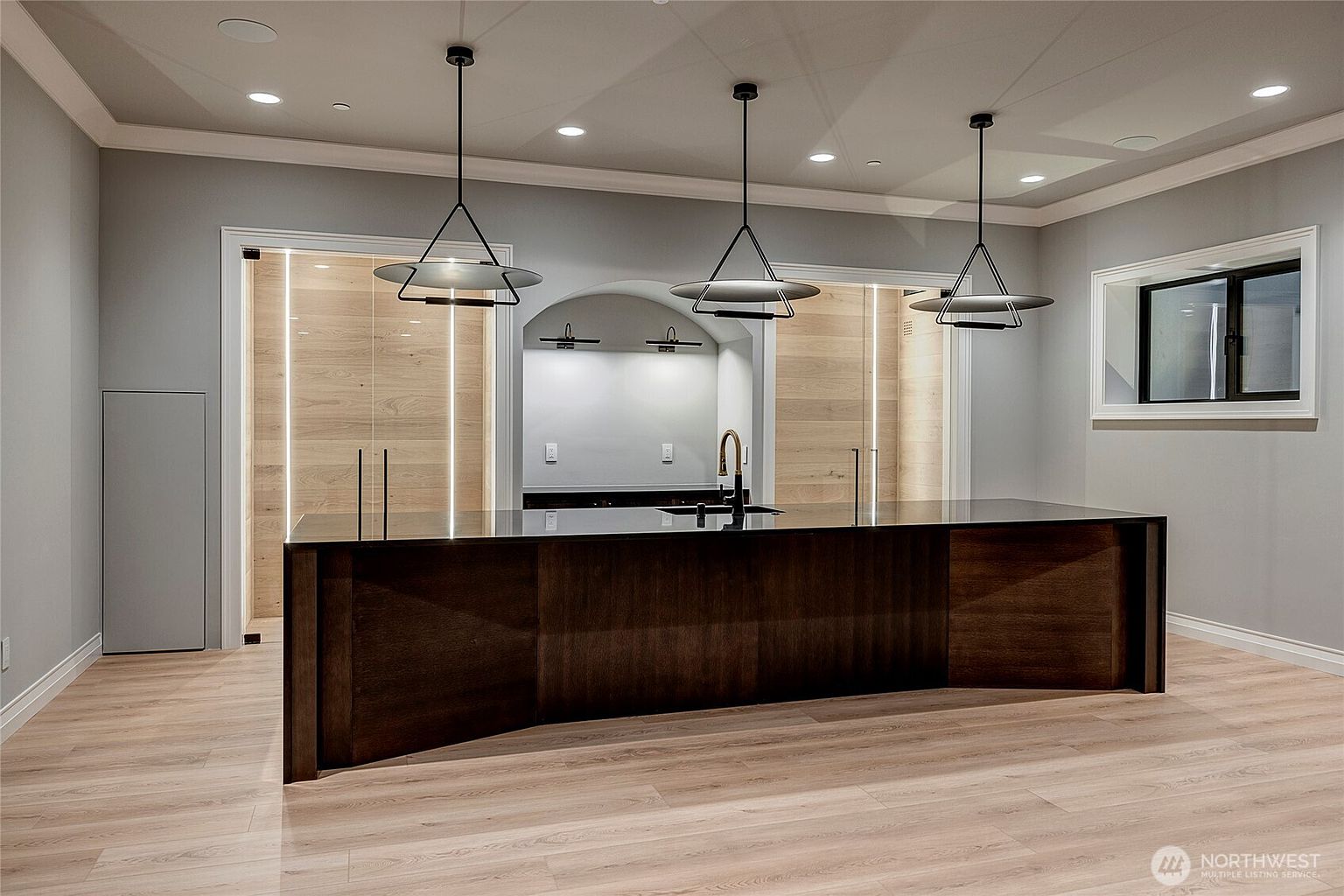 This modern kitchen features a large, dark wood island with a sleek black countertop, serving as the centerpiece of the room. Behind the island, an arched alcove with a sink and minimalist lighting creates a sophisticated focal point, flanked by light wood-paneled walls with integrated vertical LED strips. The space is illuminated by three contemporary pendant lights, contributing to a clean, high-end, and minimalist aesthetic.