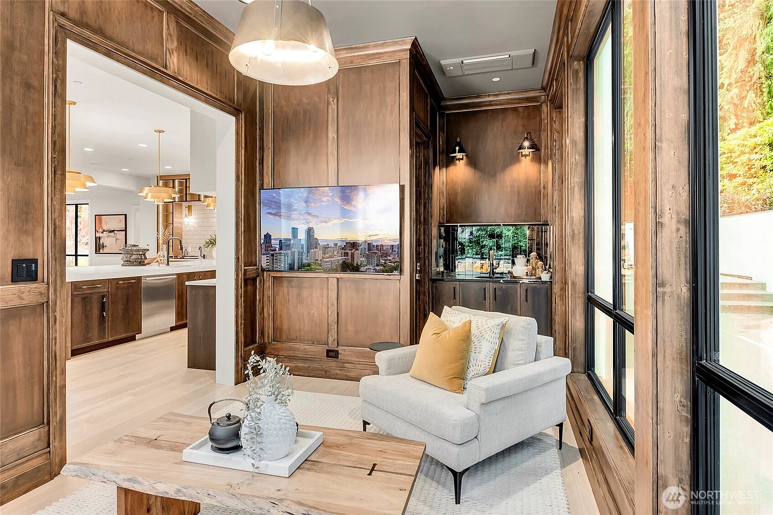 This sophisticated living space features rich, wood-paneled walls that create a warm and luxurious atmosphere. A comfortable light-gray armchair sits beside a large window, while a flat-screen television is mounted on the central wood wall above a small wet bar area. The room flows seamlessly into a bright, modern kitchen visible through the wide doorway, highlighting an open-concept design.