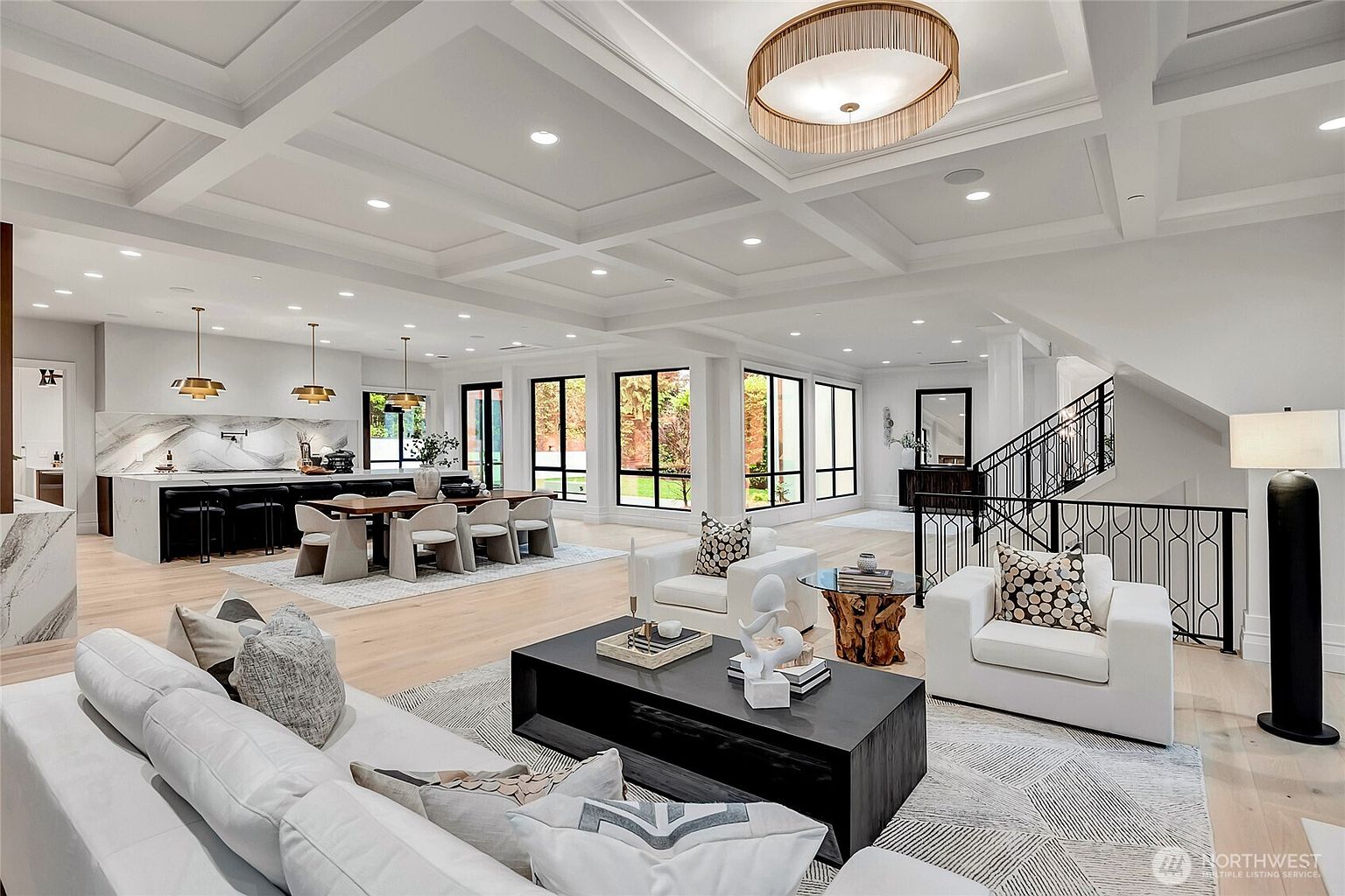 This expansive, open-concept living area features a sophisticated modern design with a coffered ceiling and light hardwood flooring. The space seamlessly integrates a formal dining area and a sleek kitchen, all anchored by a large, contemporary living room with plush white seating and a bold black coffee table. Large windows provide abundant natural light and views of the exterior, creating an airy and luxurious atmosphere perfect for entertaining.