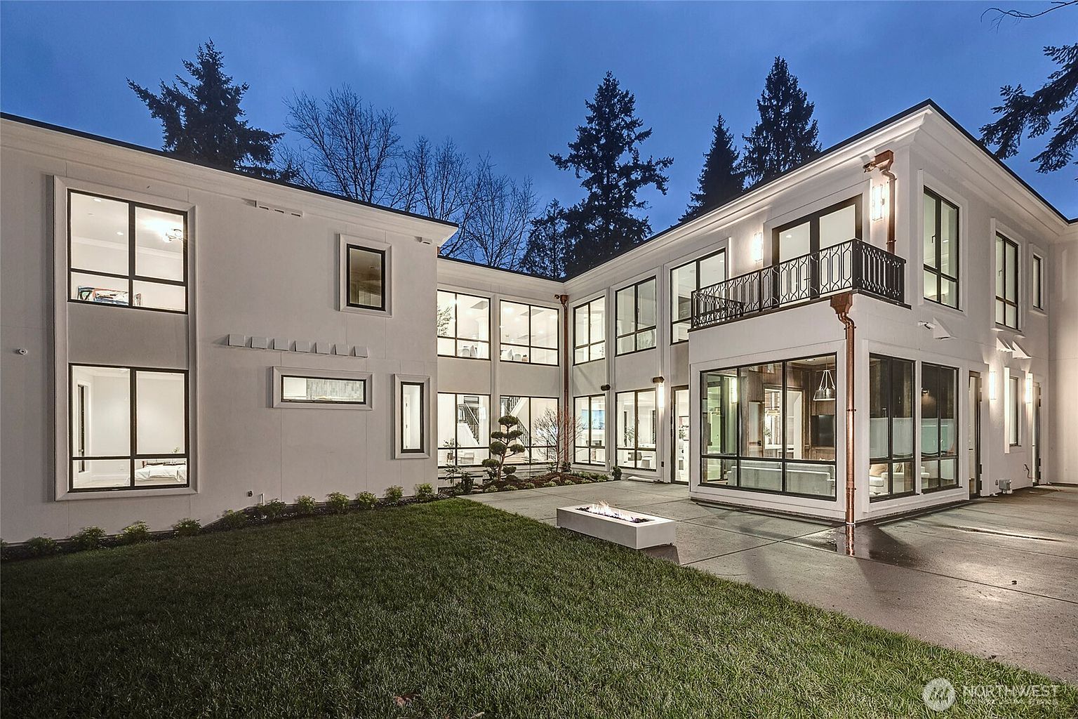 This modern, two-story luxury residence features a crisp white stucco facade with expansive floor-to-ceiling windows and a sleek, contemporary architectural design. The rear exterior includes a manicured lawn, a concrete patio with a built-in linear fire pit, and a second-story balcony with black metal railings. The twilight setting highlights the home's sophisticated lighting and clean lines, creating an inviting and upscale atmosphere.